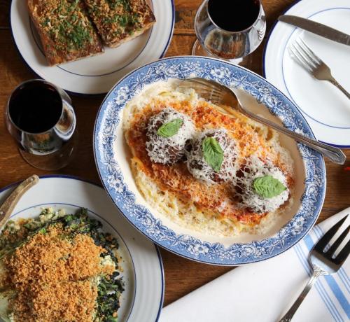 A beautifully plated dish of pasta with meatballs, topped with cheese and herbs, alongside wine and side dishes on a wooden table.