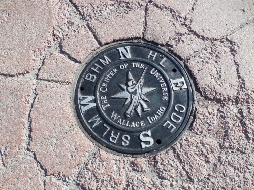 A metal marker on the ground reads "The Center of the Universe, Wallace, Idaho," surrounded by cracked pavement.