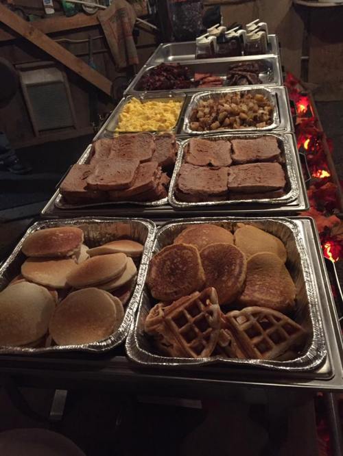 A buffet table with trays of pancakes, waffles, eggs, meats, and potatoes in a rustic setting.