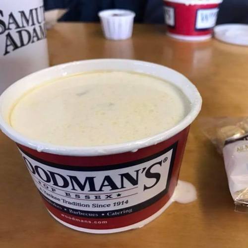 A cup of creamy clam chowder from Woodman's, with a red logo and a wooden table in the background.