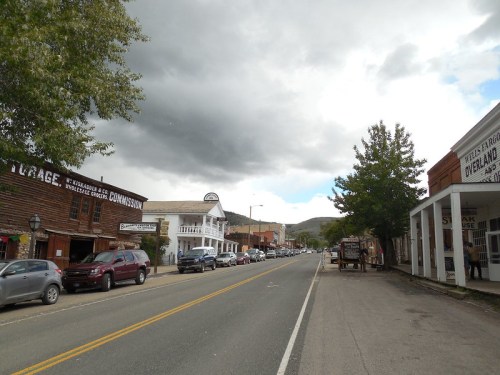 A quiet street in a small town with historic buildings, parked cars, and a cloudy sky overhead.