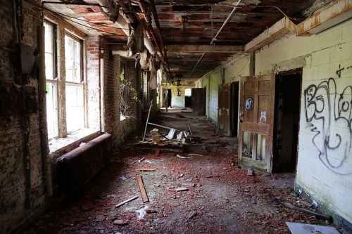 Abandoned hallway with broken windows, debris on the floor, and graffiti on the walls.
