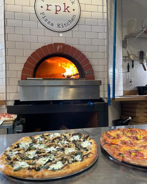 Two pizzas on a countertop in front of a wood-fired oven, with flames visible inside the oven.