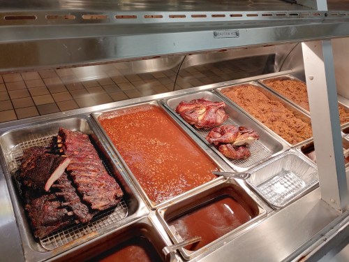 A display of barbecue dishes, including ribs, baked beans, and pulled pork in metal trays.