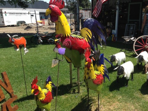 Colorful metal sculptures of roosters and sheep in a grassy area, with a fence and buildings in the background.