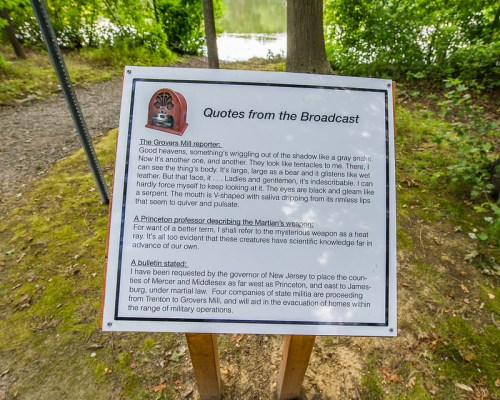 Sign with quotes about the Grovers Mill Martians and a bulletin about evacuations, set by a serene lake.