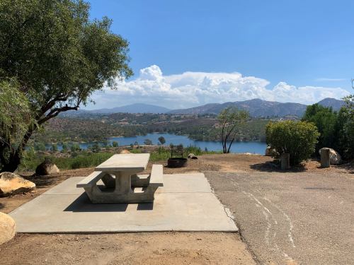 A concrete picnic table overlooks a serene lake surrounded by hills and trees under a clear blue sky.