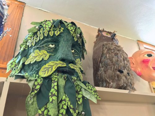 A green, leafy mask resembling a face, next to an owl figure and a large, colorful mask on a shelf.