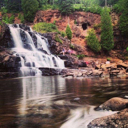 A serene waterfall cascading over rocks into a calm pool, surrounded by lush greenery and rugged cliffs.