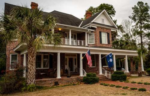 A large brick house with a porch, featuring American and South Carolina flags, surrounded by palm trees and greenery.