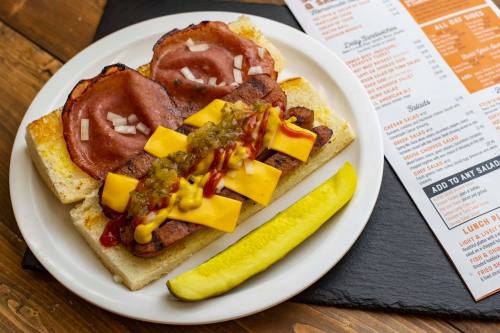 A plate with a sandwich featuring sliced meats, cheese, and condiments, alongside a pickle and a menu.