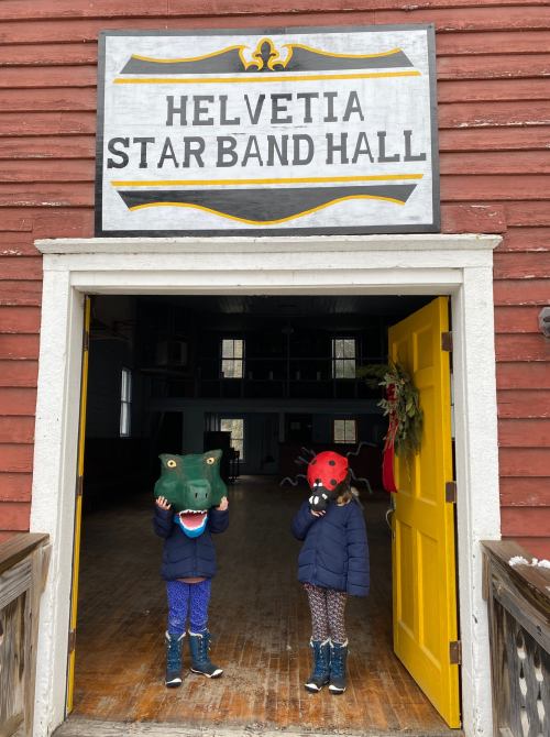 Two children wearing animal masks stand at the entrance of Helvetia Star Band Hall, with a festive wreath nearby.