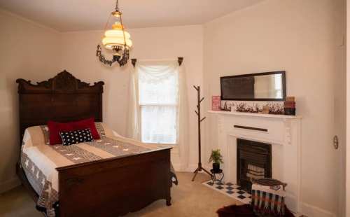 Cozy bedroom featuring a wooden bed, decorative quilt, fireplace, and a window with soft curtains.