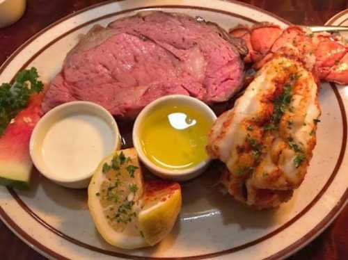 A plate featuring prime rib, lobster tail, lemon, dipping sauces, and a slice of watermelon.