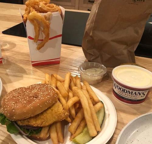 A plate with a fried chicken sandwich, fries, a pickle, and a container of coleslaw, alongside a box of onion rings.