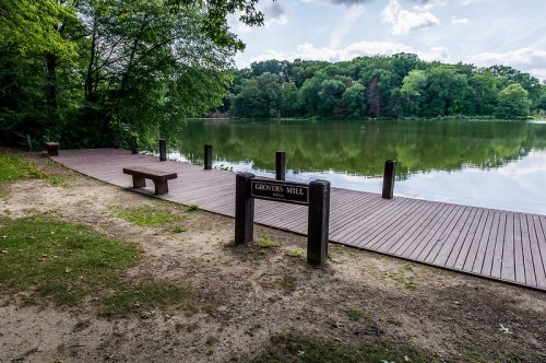 A serene lakeside view at Grovers Mill, featuring a wooden dock and lush green trees under a cloudy sky.