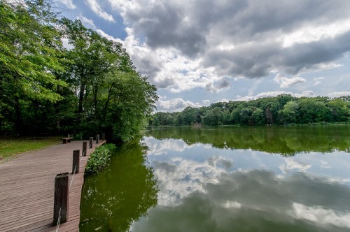 A serene lake surrounded by trees, reflecting clouds and greenery, with a wooden path along the water's edge.