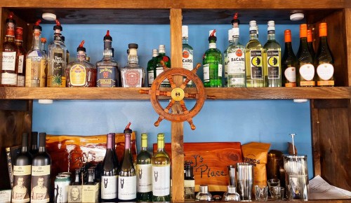 A wooden shelf displaying various bottles of liquor, wine, and mixers, with a ship's wheel in the center.