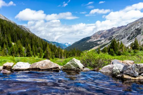A serene mountain landscape with a clear pool of water in the foreground, surrounded by lush greenery and distant peaks.