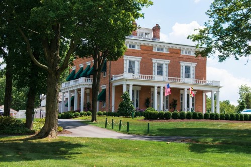 A large brick mansion with white columns, surrounded by green lawns and trees, featuring American and Maryland flags.