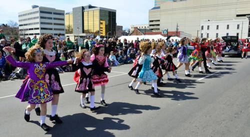 A group of young dancers in colorful costumes performing at a parade, with spectators watching in the background.
