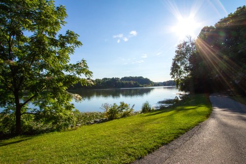 A serene lakeside view with sunlight reflecting on the water, surrounded by trees and a walking path.