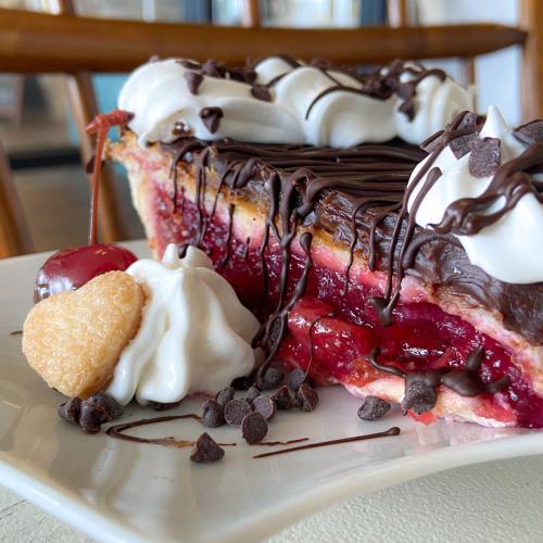 A slice of chocolate-covered cherry pie topped with whipped cream, chocolate chips, and a heart-shaped cookie.