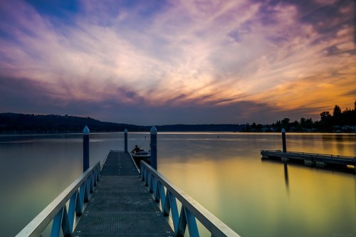 A serene lake at sunset, with a dock extending into calm waters and colorful clouds reflecting in the sky.