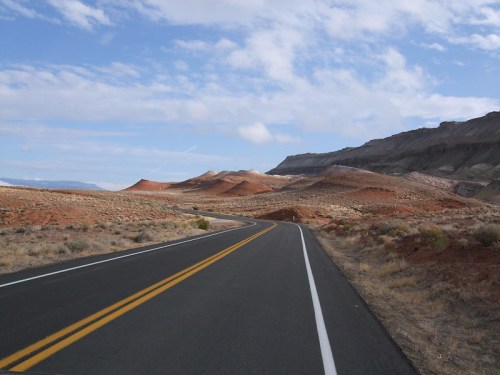 A winding road through a desert landscape with red hills and a blue sky dotted with clouds.