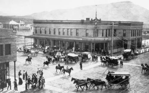 Historic black-and-white photo of a bustling town square with horse-drawn carriages and people in period clothing.