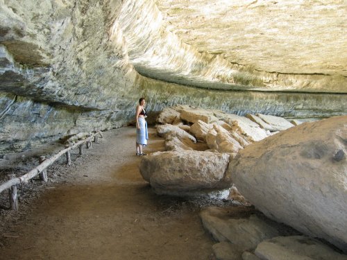 A person stands inside a large cave, surrounded by rocky formations and a dirt path. Natural light illuminates the space.