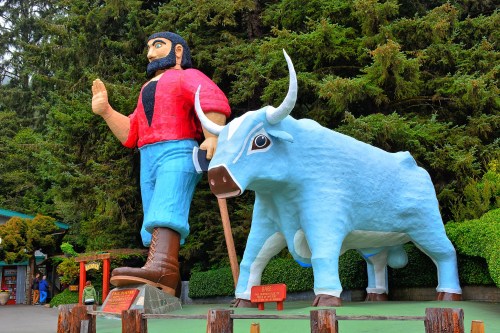 A large statue of a man in a red shirt and blue pants stands next to a blue cow statue, surrounded by greenery.