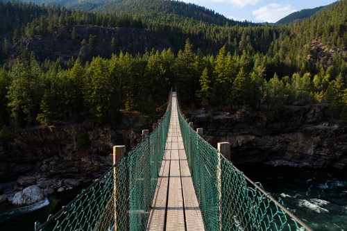 A suspension bridge stretches over a river, surrounded by lush green trees and mountains under a clear blue sky.