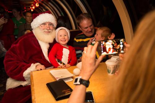 A child in a Santa outfit smiles with Santa Claus at a festive table, while adults take photos in a cozy setting.