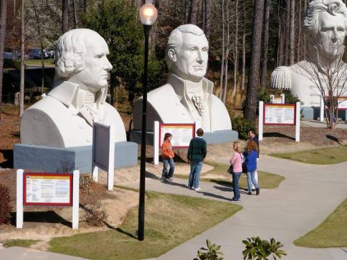 Visitors stand near large stone busts of historical figures in a park setting, surrounded by trees and informational signs.
