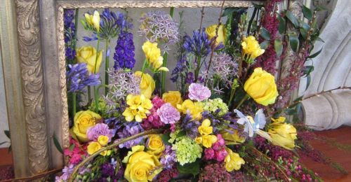 A vibrant floral arrangement featuring yellow roses, purple blooms, and greenery, framed in an ornate gold frame.