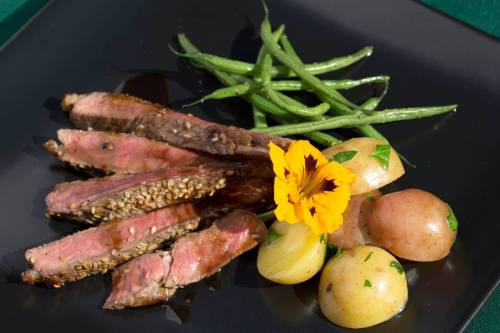 A plate featuring sliced steak, green beans, and baby potatoes, garnished with a yellow flower.