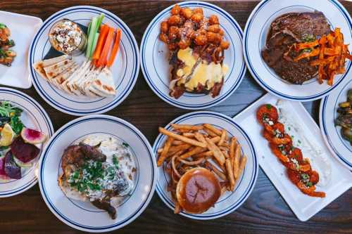 A variety of dishes including salads, fries, tater tots, grilled meats, and colorful vegetables on a wooden table.