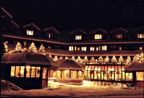 A cozy hotel illuminated with festive lights, surrounded by snow-covered trees at night.