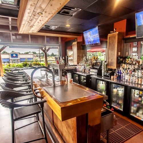 A modern bar with a wooden counter, shelves of liquor, and bar stools, featuring large windows and TVs in the background.