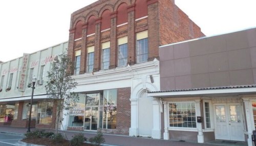 Historic brick building with arched windows, flanked by modern storefronts, in a small town setting.