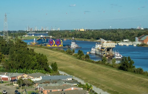 A river scene with tugboats and large machinery, surrounded by greenery and distant city skyline under a clear blue sky.