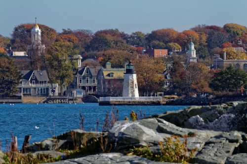 A lighthouse stands on a rocky shore, with colorful autumn trees and historic buildings in the background by the water.