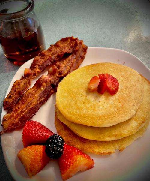 A plate with three pancakes topped with a strawberry, crispy bacon, and fresh berries, alongside a jar of syrup.