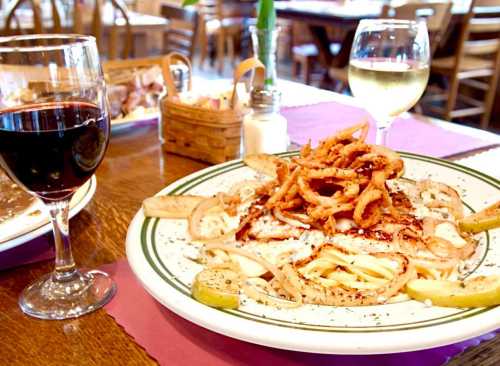 A plate of pasta topped with fried onions, served with a glass of red wine and a glass of white wine in a restaurant setting.