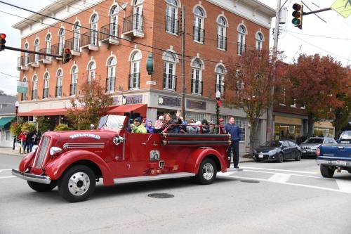A vintage red fire truck drives through a town, filled with smiling passengers, in front of brick buildings and shops.