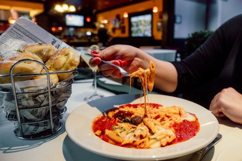 A hand holds a fork with cheesy pasta over a plate, accompanied by a basket of breadsticks in a restaurant setting.
