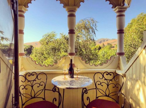 A charming balcony with a round table, two glasses, and a bottle, surrounded by trees and mountains under a clear blue sky.