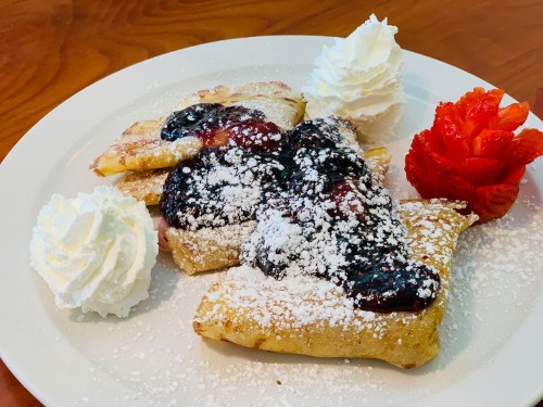 A plate of crepes topped with berry sauce, powdered sugar, whipped cream, and a strawberry garnish.