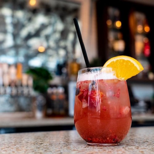 A refreshing cocktail with berries and an orange slice, served in a glass with a straw on a bar countertop.